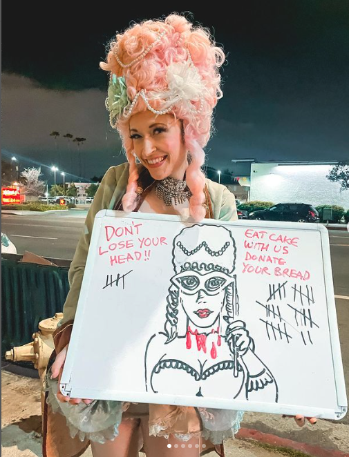 Picketer dressed as Marie Antoinette, with a white board sign that says 'Don't lose your head! Eat cake with us, donate your bread' with a drawing of Marie Antoinette with blood dripping from her neck, and tally marks showing the number 32