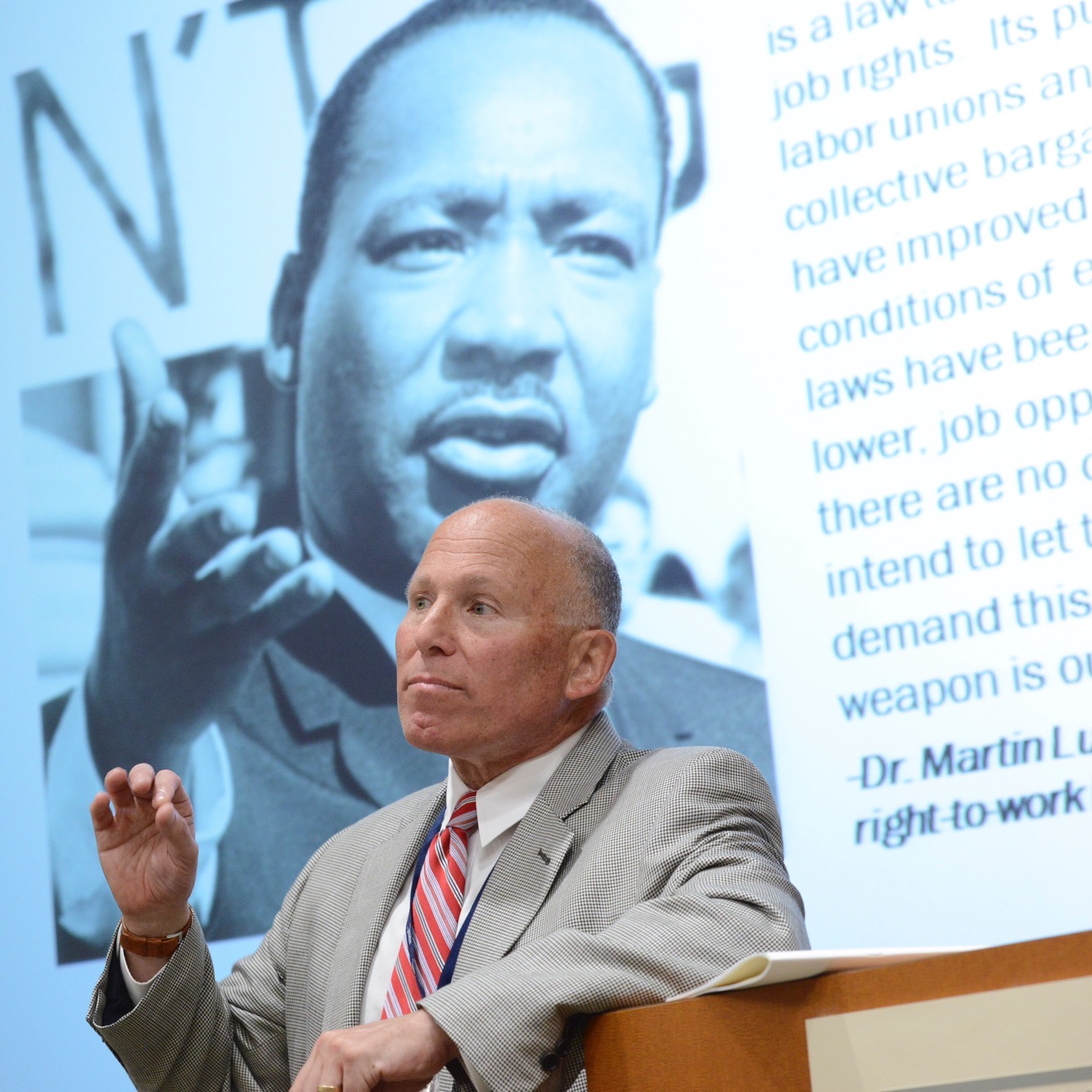 Photo of man in suit, elbow on lectern, mid-speech. Projected behind him is an image of Rev. Dr. Martin Luther King, Jr.