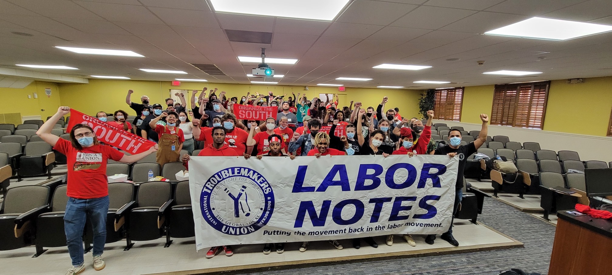 Photo from Alabama Troublemakers School 2022 shows a crowd of people in an auditorium grouped behind a Labor Notes banner, fists raised.