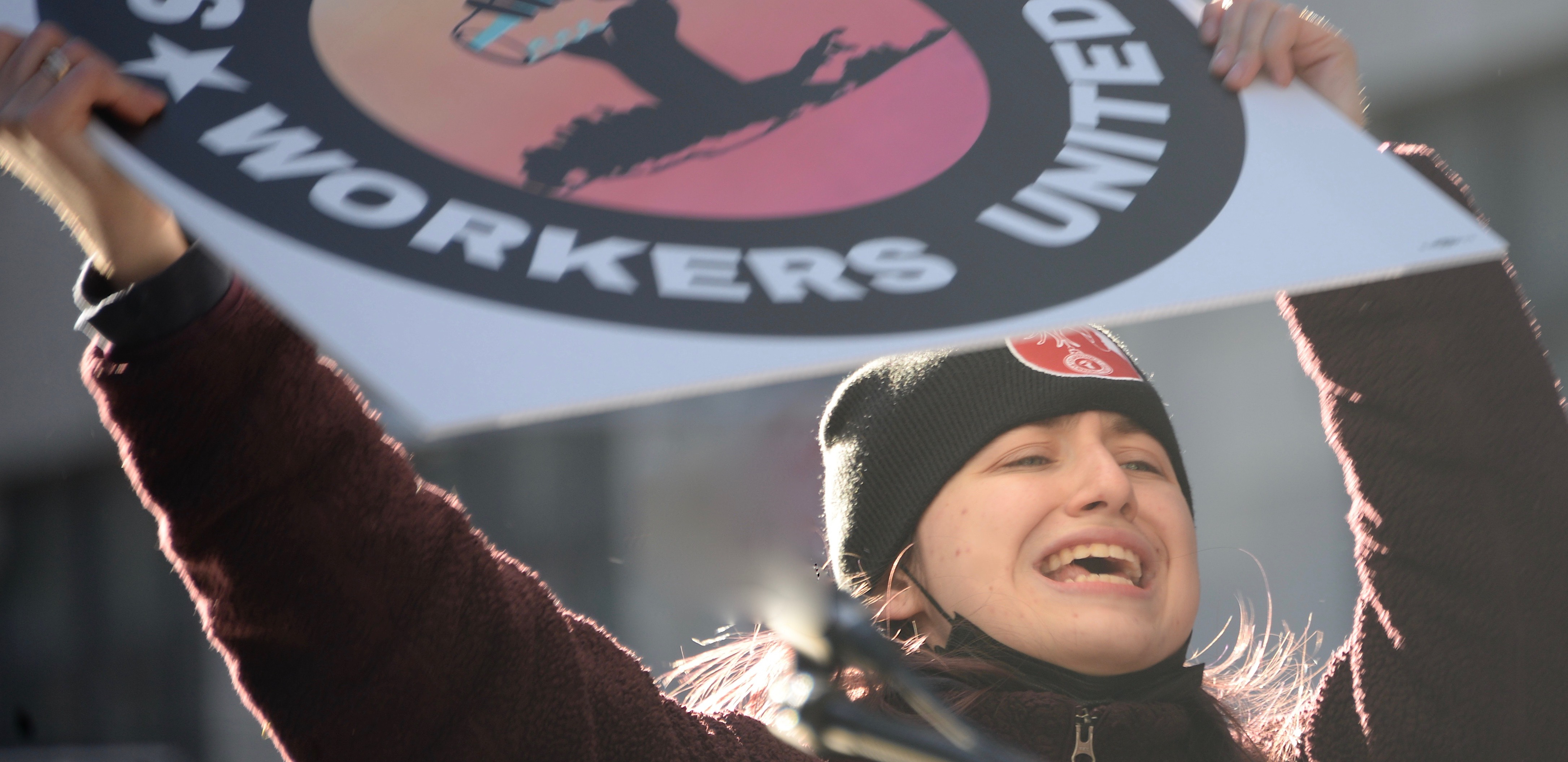 Starbucks Woman in a warm hat holds Starbucks Workers United sign high with sun behind her