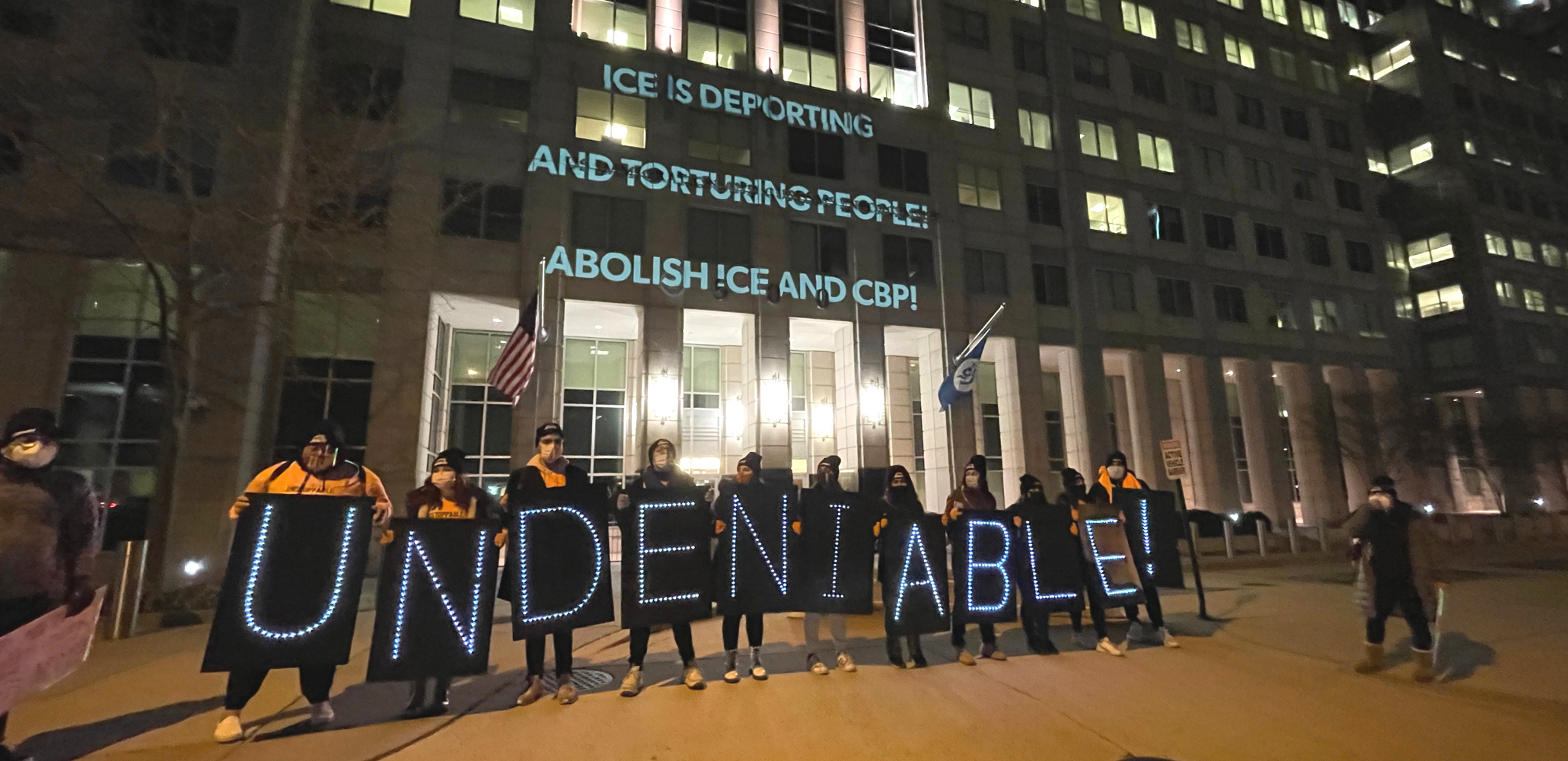 People holding lit signs spelling out "UNDENIABLE" stand in front of a building. On the building are projected the words: "ICE IS DEPORTING AND TORTURING PEOPLE! ABOLISH ICE AND CBP!"