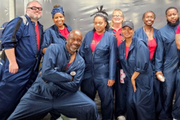 Nine workers (most of them Black, a mix of women and men) pose in red T-shirts and blue work overalls, apparently inside the plant. Their pose evinces camaraderie and confidence. One man in the front has a particularly dynamic pose with a lunge, crossed arms, and a big smile.