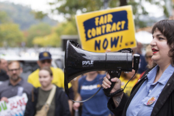 A woman with long dark hair speaks into a megaphone to a crowd that is visible beyond her, one holding a “Fair Contract Now” sign.
