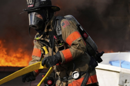 A firefighter in full protective equipment moves a yellow hose with fire in the background
