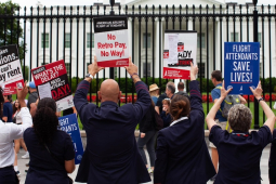 A line of seven flight attendants in uniform, viewed from behind, stands at the White House gates holding up various printed signs. Messages say: "AA makes billions, we can't pay rent." "Robert, what's the delay? We need a contract today!" "American Airlines flight attendants: No retro pay, no way!" "Flight attendants save lives!" "Corporate greed doesn't fly." "Trying to get pAAid." It appears the group includes men and women, and most are people of color.