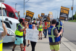 Workers in high-visibility vests picket with ‘Amazon workers on strike’ signs picket by a road, one handmade sign reads ‘Stop the captive audience’