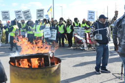 In the foreground a burn barrel, one striker walking purposefully, one raising a fist. In the background a large crowd of strikers carrying "CUPW on strike" picket signs and wearing safety-yellow vests.