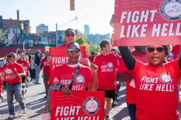 Black and white women and men march through a Detroit street. Everyone is wearing a red "fight like hell" T-shirt with the NALC logo, and some people are carrying printed signs with the same design. Most prominent in the frame, looking directly into the camera, are two Black women in sunglasses, one raising a fist at shoulder level and the other holding a sign above her head. 