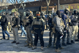Agents covered head to toe in tactical gear and warm clothing stand in a diamond formation, facing outward, in a city street. Their faces are covered. The vests say "police" though they are not in uniform. They all wear heavy boots and some carry spray cans. Visible behind them is a one-story brick building lined with retail shops, with faded signs reading "8 Street Nourish" and "Tobacco & Mas."
