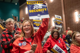 People, mostly wearing red, hoist printed signs in a packed indoor rally. A woman in the foreground, smiling, wears a "no tiers" button. Her sign, held high, reads "UAW: Fight for Job Security." Other visible signs say "UAW: Fight for No Tiers" and "UAW: It's Our Time at Local 2093 American Axle." A man behind her, applauding, wears a red and black buffalo plaid shirt. Most people visible in the photo appear to be white.