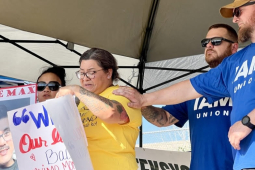 A woman cries while two men in blue Machinists t-shirts comfort her with a hand on her shoulder. Next to her is a woman holding a sign that says “Free Max.”