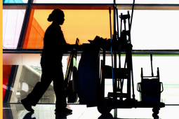 A woman custodian pushes a cart full of implements in profile past colorful windows.