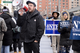 A line of men and women picketers with NTEU signs march on the right while a man with a bullhorn chants on the right