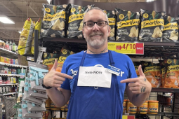 A man in in front of grocery shelves points to a printed-out “Vote No” sign pinned to his blue shop apron.