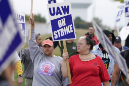 The camera focuses on two women in a crowd of picketers, diverse in race and gender, carrying printed blue and white "UAW on strike" picket signs. The women are wearing different UAW T-shirts. One has the collar cut off, revealing part of a tattoo that reads, in cursive, "darkness light shall prevail." Both women wear serious, determined expressions.