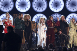 A row of musicians with large clocks behind them perform, with Dolly Parton in the center