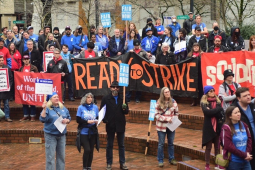 People rally on brick steps (Portland's Pioneer Courthouse Squre) on a rainy day. Many wear blue or red T-shirts under their coats. Banners say "Ready to strike" and "Soldarity." Picket signs say "Fair Contract Now," "Solidarity with Strike-Ready Teachers," and "Essential Wages for Essential Workers."