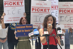 A woman speaks into a microphone outside while others stand behind her with signs that say “Picket ICE”