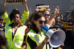 Several workers in green high-viz vests chant and hold signs, one woman in front speaks into a megaphone.