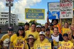 Nurses in yellow shirts pose smiling. Most are women; most appear Native Hawaiian or Asian. Many wear ballcaps. Some carry printed signs saying "ULP strike," "Zero tolerance for retaliation," or "on strike for better patient care." Two carry handmade signs--one with an image of a duck and the words "Unsafe staffing leads to quack-cidents," and the other with the logo of the show "Friends" crossed out, the word "Bullies" written in the same style, followed by "Episode 2: The One Where HPH Locks Us Out."