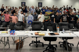 A group of 50 people stand behind a bargaining table with fists raised. 
