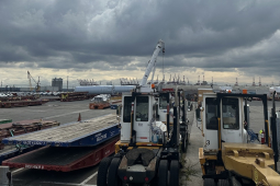 Small port vehicles are in the foreground, and harbor cranes are visible in the distance.