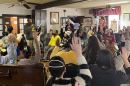 A group of people in chairs crowd a room listening to a speaker.