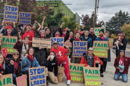 A group of workers and supporters hold cardboard signs reading Stop Unfair Labor Practices and Fair Contract Now. Many are wearing red bandanas.