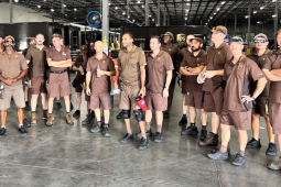 Fifteen workers in brown UPS uniforms stand in a semicircle while another uniformed worker speaks to them