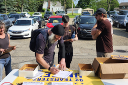 A group of Teamsters in a parking lot stand around a table with petitions and leaflets on it. One is filling out a sign-up sheet.