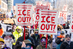 View of a section of a giant crowd marching, on a bright sunny day, bundled against extreme cold. Prominent picket signs that look screen-printed or stenciled say "Neighbors say ICE out!" in crisp red on white background.