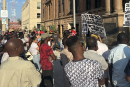 A group of workers, mostly Black, face an official-looking building with signs.
