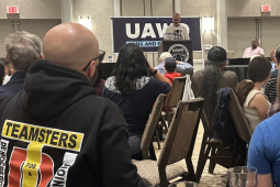 An audience listens to a man at a lectern. One audience member has a blue shirt with “Union Strong” on the back.