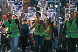Strikers march outdoors. The three in front, wearing neon vests of picket captains, are chanting energetically, two with fists in air, one holding bullhorn. Crowd behind them holds "UAW on strike" picket signs.