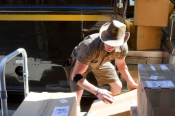 A uniformed UPS delivery driver, wearing a UPS-branded cowboy hat, lifts large packages onto a rolling cart in front of his delivery truck. More large packages are visible through the truck's open door.