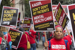 Strikers in red march with printed picket signs. In the center is a Black woman with a serious expression. Her sign reads "Kaiser, don't deny my patients mental health care. KaiserDontDeny.org."