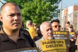 Crowd in UPS brown uniforms stands outdoors looking resolute, some holding signs. One sign visible reads "Make UPS Deliver!"
