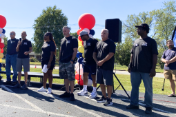 About a dozen adults and a couple children, white and Black, men and women, stand mostly silhouetted in bright sun. Most wear black union T-shirts.They're outdoors, on pavement in front of grass. One holds a mic. Some hold printed signs that say, "IUE-CWA, Better Wages." There are also two columns of balloons, each a spiral design in white, red, yellow, and black.