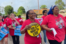 Women, mostly Black women, march outdoors in red T-shirts that say "Organize" with a telephone logo. Most prominent in the foreground are three women who have linked elbows and are smiling big. The woman on the left has a yellow round sign that says "Everybody's got a right to live." The woman in the middle has blue hair. The woman on the right is holding a light blue sign we can't see, but it probably matches signs visible behind which say "CWA on strike against Maximus." One woman is smoking.