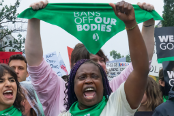 Women shout and raise their fists at a rally. Many are wearing green bandannas and one is holding the bandanna up in the air: it says "Bans off our bodies." A sign held up in background says "We won't go back."