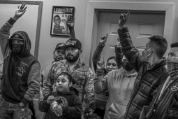 Black and white photo shows workers raising their hands, some holding kids, indoors with a small Rosie the Riveter poster on a nearby wall.