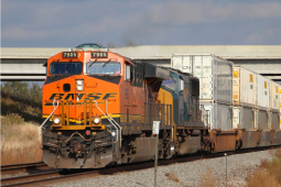 A freight train passes along a track under an overpass. The orange engine is labeled BNSF. It is pulling many white freight cars.