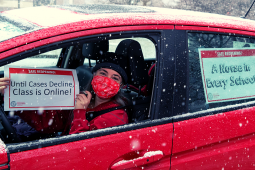 Person in driver's seat of red car, wearing red mask, holds sign: "Safe reopening! Until cases decline, class is online!" Taped to back window of car is another sign: "A nurse in every school!" It is snowing.