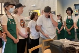 Young workers in Starbucks aprons and SBWU logo shirts stand in a semicircle, all in calm postures mostly with hands clasped at rest in front of them. In the center is the manager in a white shirt, talking into a cell phone.