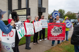 People with signs stand on grass outside an office building. The man speaking holds a red banner that says, "Solo organizados conquistaremos nuestros derechos laborales. Casa Bajia Obrera." Others listening hold printed blue UAW logo signs and hand-written signs that say "Shame on VU" or "End the blacklist." 