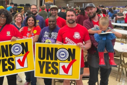Ten smiling people pose together in a crowded meeting room. Two are holding huge yellow "Union Yes" signs with the UAW logo. Most are wearing red, blue, or purple T-shirts with UAW logo or organizing-themed slogans. The man on the far right holds a small child in his arms.