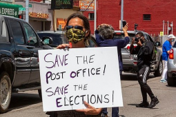 A masked protester holds a sign: "Save the Post Office! Save the Elections!" Behind the person, traffic and a "Coney Island" (diner) across the street
