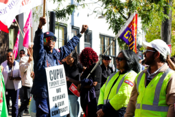 A Black worker in postal uniform, wearing reflective shades and a sandwich board sign that says "CUPW Toronto Local, We demand a fair contract," raises one fist in the air and a flag on a stick in the other hand. The flag is only partially visible but includes the word "Justice." This person is standing up on something we can't see, a step or box. Smiling people around them are holding various banners and signs, some wear safety yellow. Variety in race and gender. In sunlight dappled by shade of tree. 