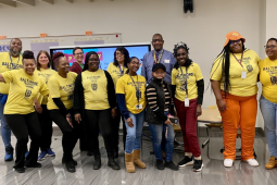 Fourteen workers, mostly Black and mostly women, pose smiling in a school classroom. Most are wearing yellow "Baltimore Teachers Union" T-shirts.