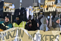 A crowd of workers stands outdoors in dim light. A smiling person with raised fist is one of several holding a handmade banner--the part visible says "Amazon Workers." Many people in crowd hold printed Teamster-logo picket signs that say: "Amazon: Obey the Law." Everyone is wearing coats with big hoods or warm hats; it looks cold out. Someone in lower left has a Staten Island T-shirt and keffiyeh